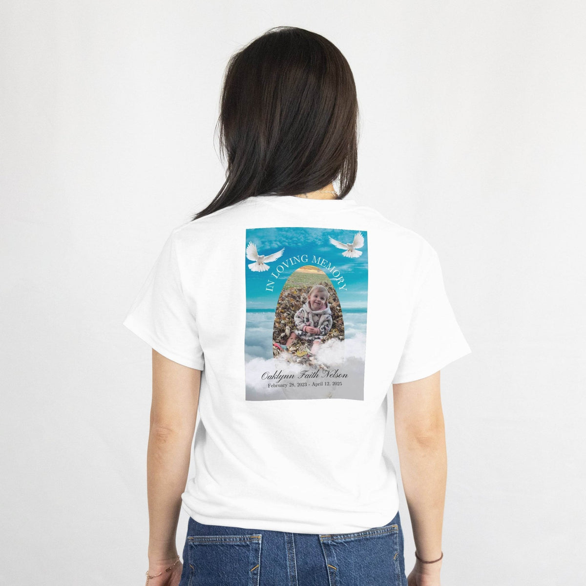 Back of a white T-shirt featuring a memorial print with a baby photo, name, dates, doves, and In Loving Memory text.