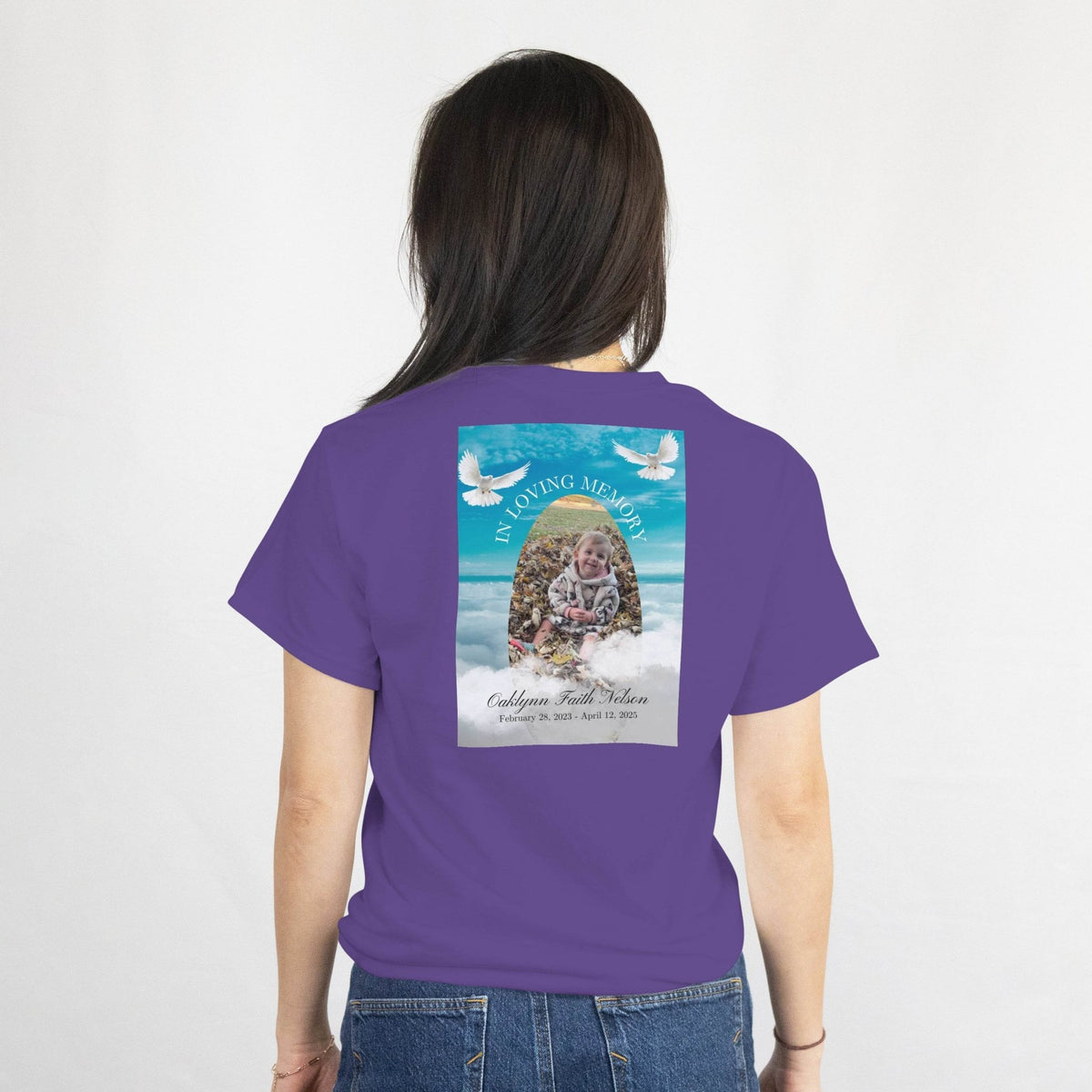 Memorial design on a purple t-shirt back featuring a baby photo in an arch, two doves, blue sky and waves, with name and dates.