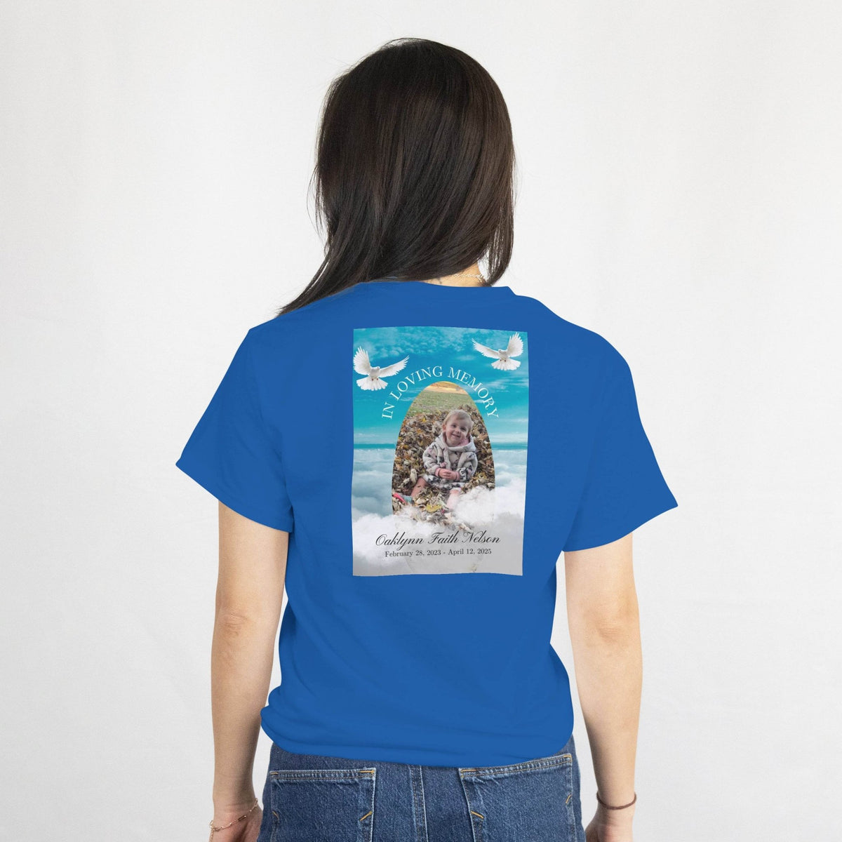 Back view of a blue memorial T-shirt with gentle doves, In Loving Memory, and a child’s photo in a frame with dates.