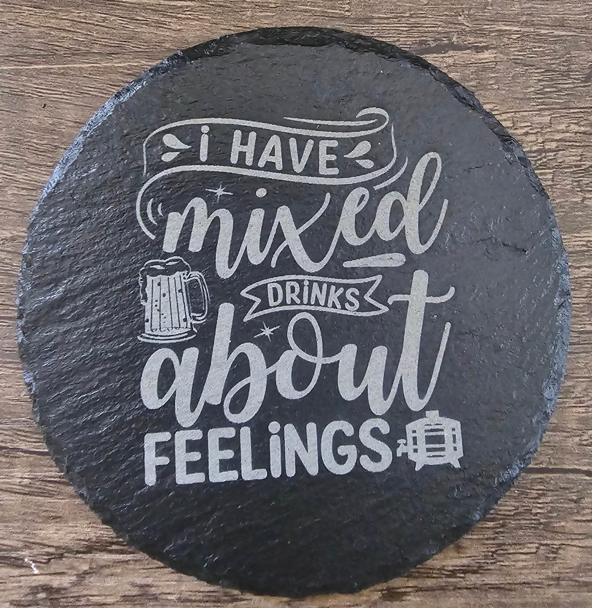 Round black slate coaster with white chalk-style typography reading I have mixed drinks about feelings.