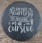 Round black slate coaster with white script I'm not drinking, I'm speaking in cursive and a small glass icon.