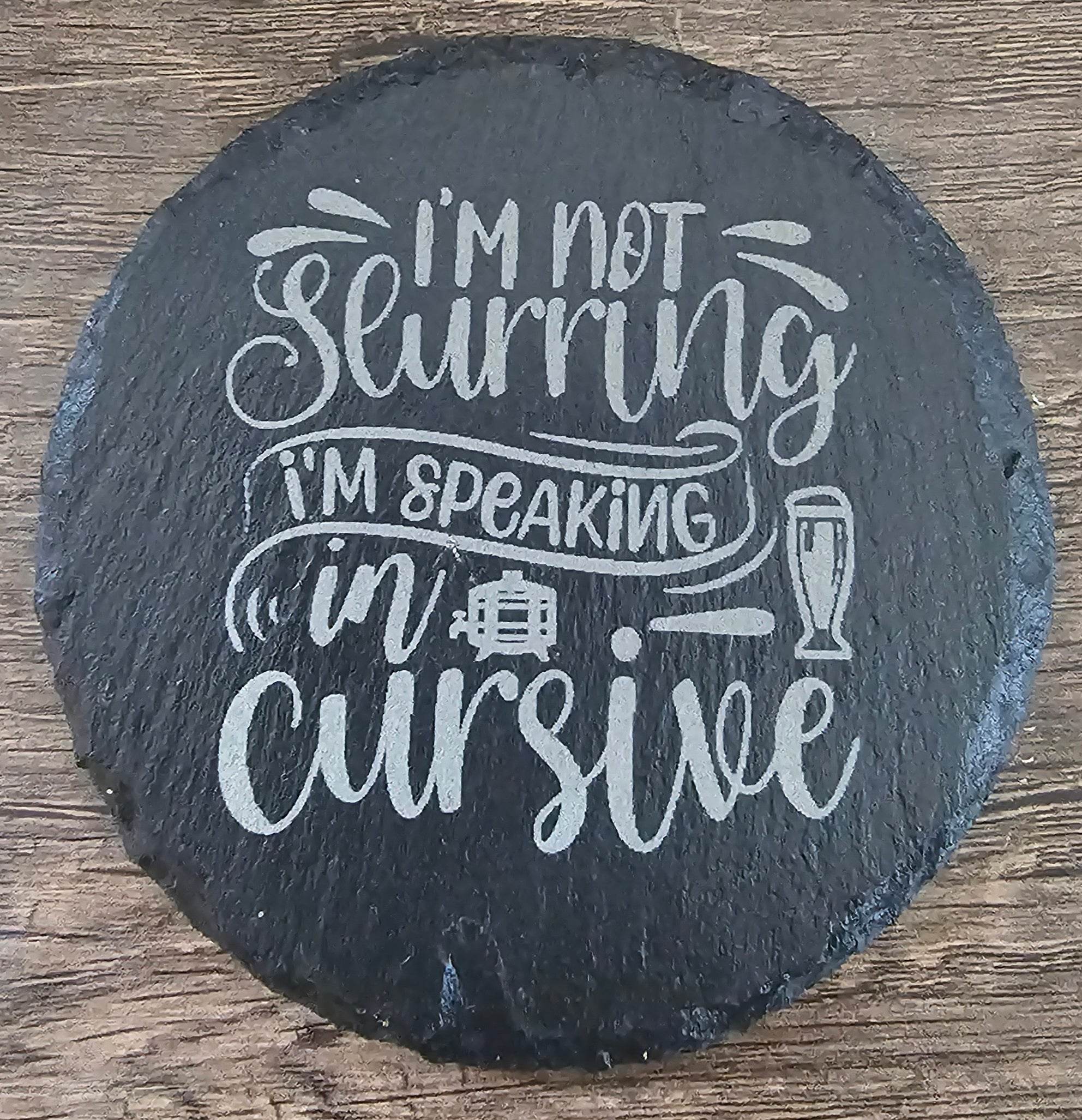 Round black slate coaster with white script I'm not drinking, I'm speaking in cursive and a small glass icon.