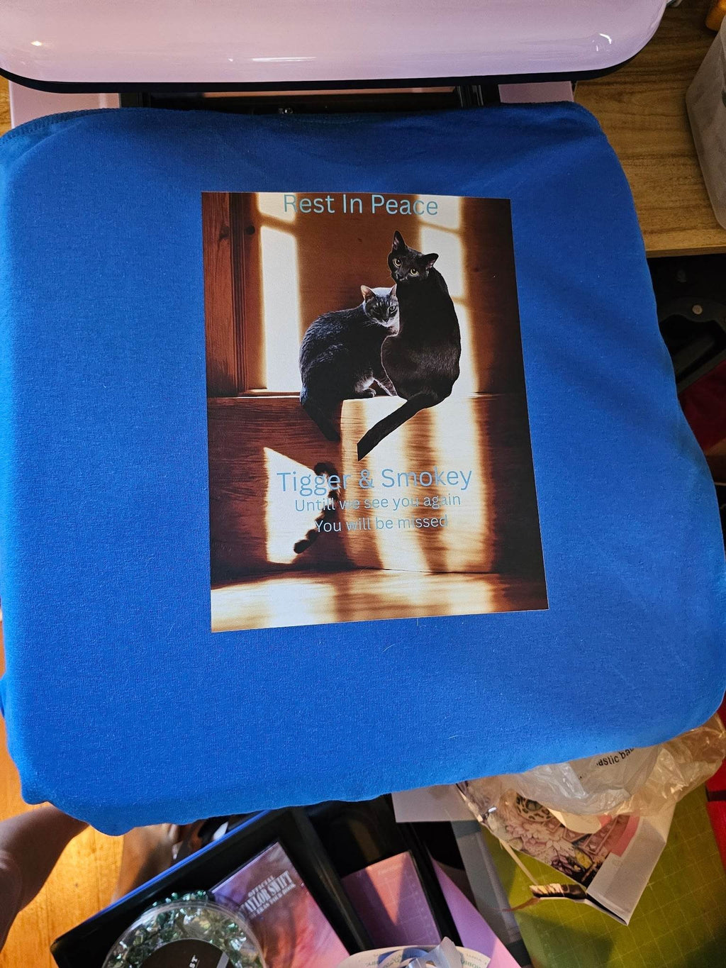 Blue memorial T-shirt laid flat featuring a print of two cats and the text Rest In Peace.