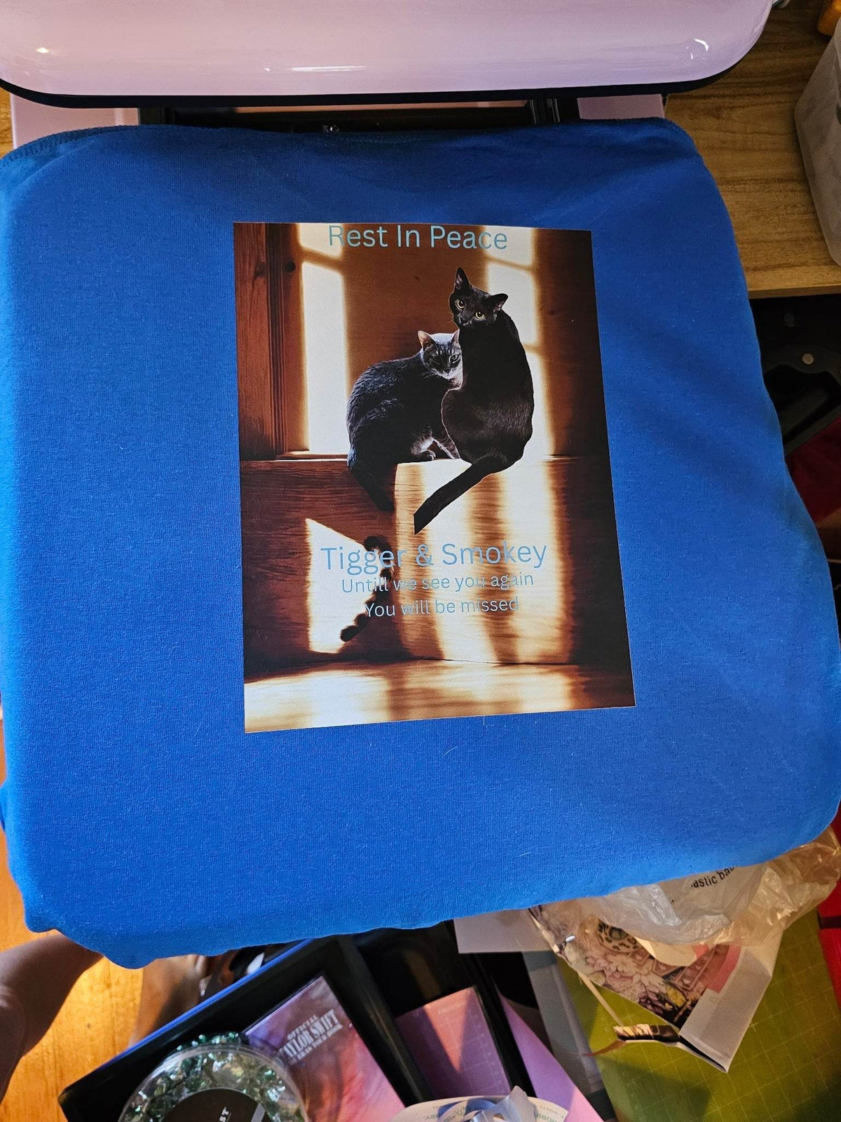 Blue memorial T-shirt laid flat featuring a print of two cats and the text Rest In Peace.