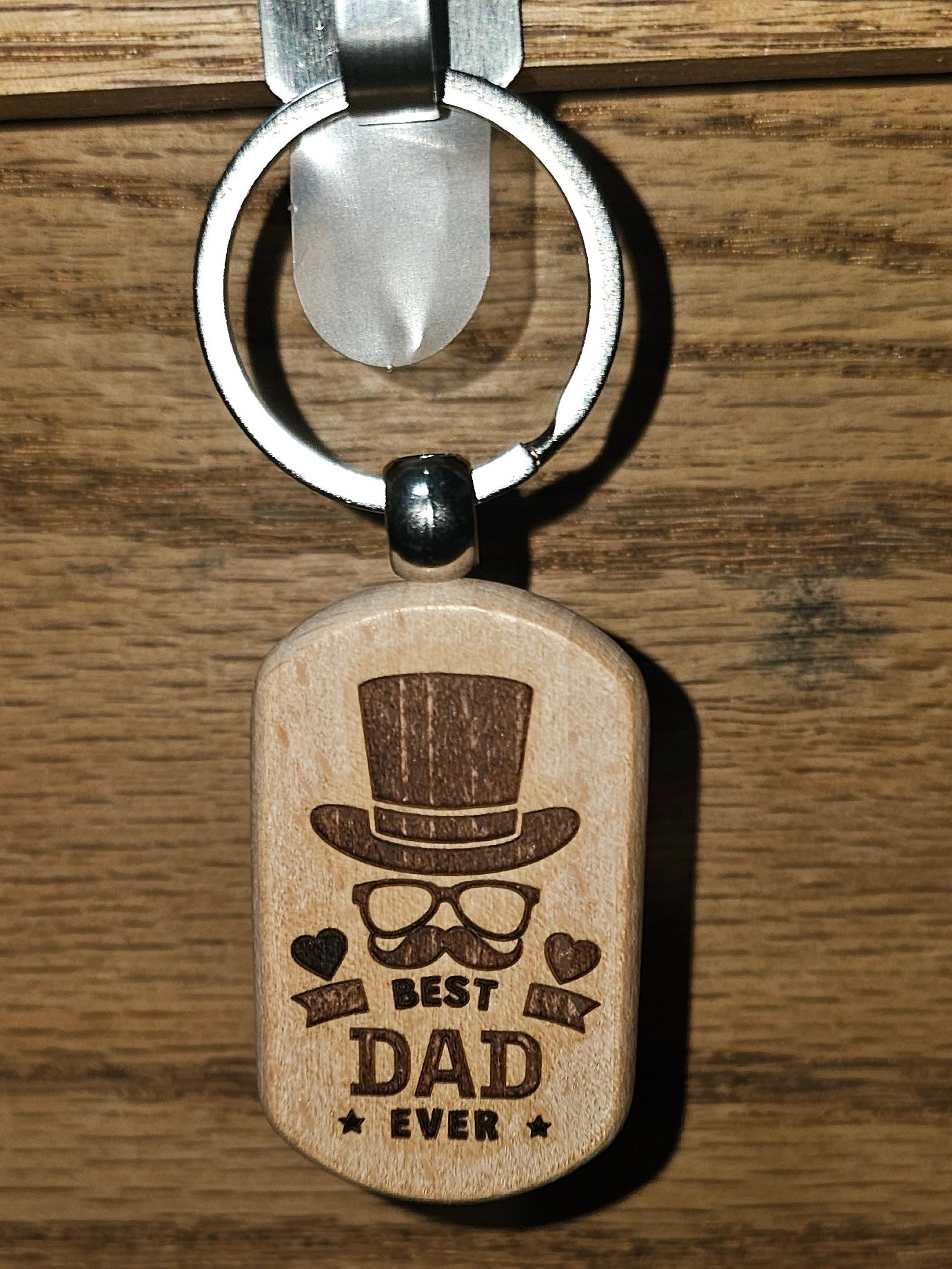 A wooden dog tag keychain with an engraved 'Best Dad Ever' design featuring a top hat, glasses, and mustache, hanging from a metal hook on a wooden surface.