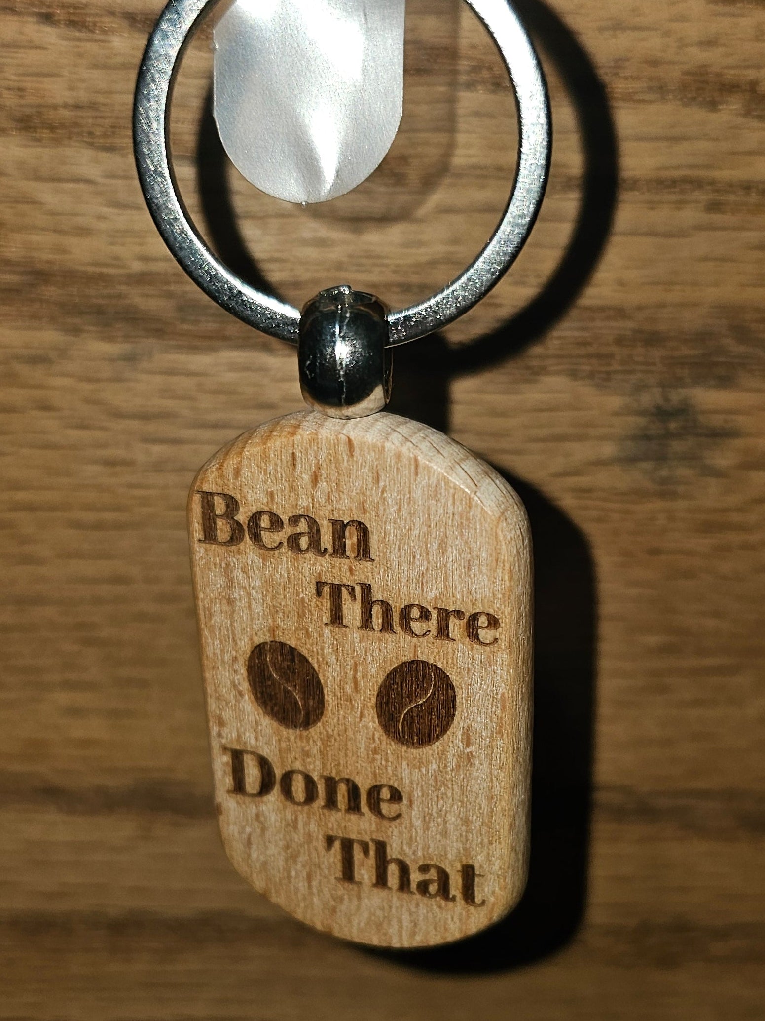 A wooden engraved keychain tag hanging from a metal ring on a wooden door. The tag is light brown wood with dark brown engraved text reading 'Bean There Done That' and two small bean illustrations.