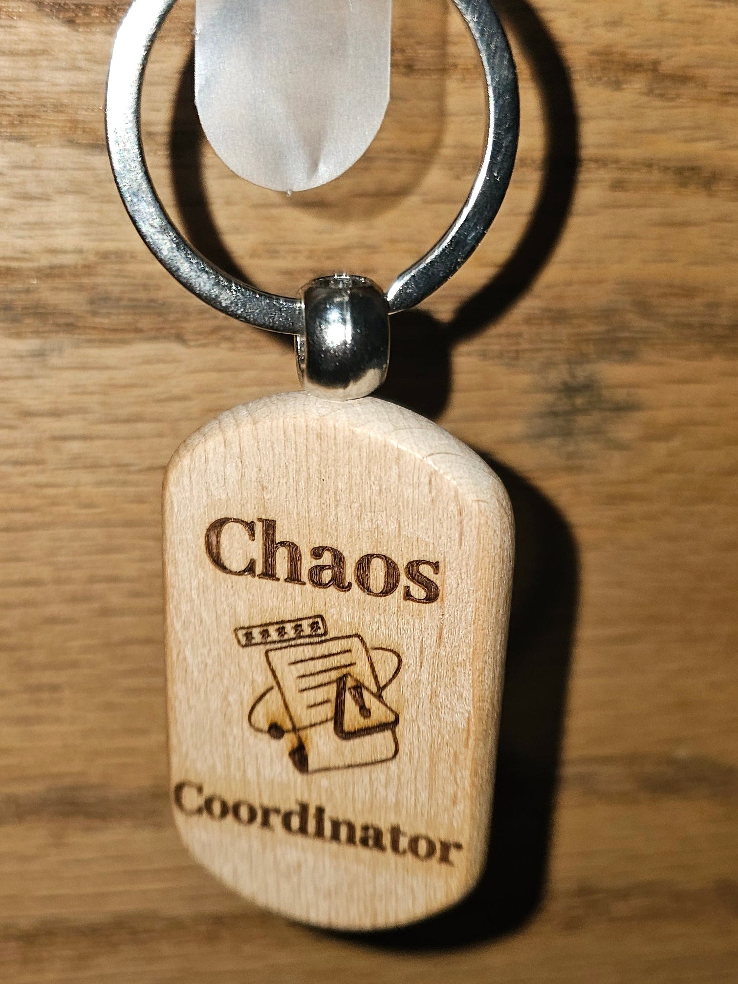 A wooden dog tag keychain engraved with 'Chaos Coordinator' and a small document icon, hanging from a metal ring on a wooden door.
