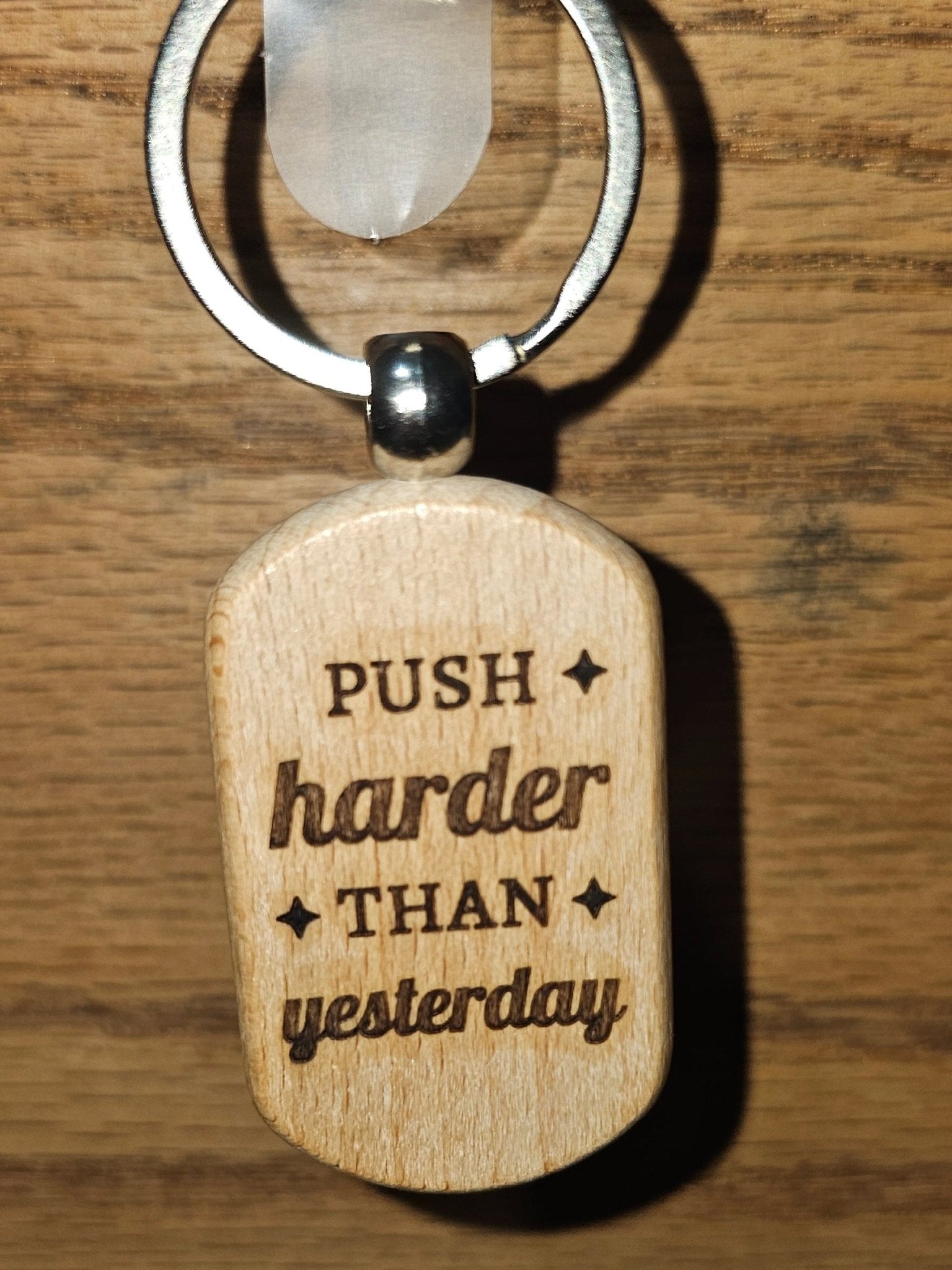 A small oval wooden keychain tag engraved with the motivational phrase 'PUSH harder THAN yesterday' in dark lettering, attached to a silver metal split ring and key blade, resting on a brown wooden surface.