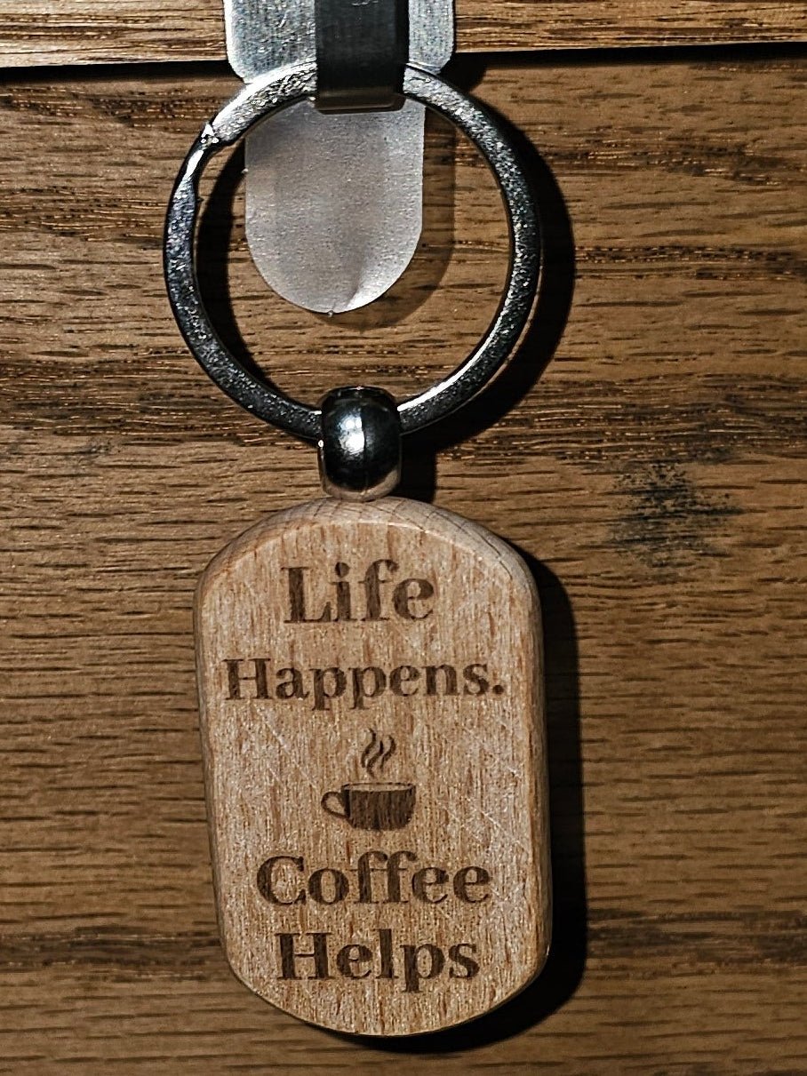 A wooden dog-tag style keychain with engraved text reading 'Life Happens Coffee Helps' hanging from a black ring on a metal hook attached to a wooden surface.