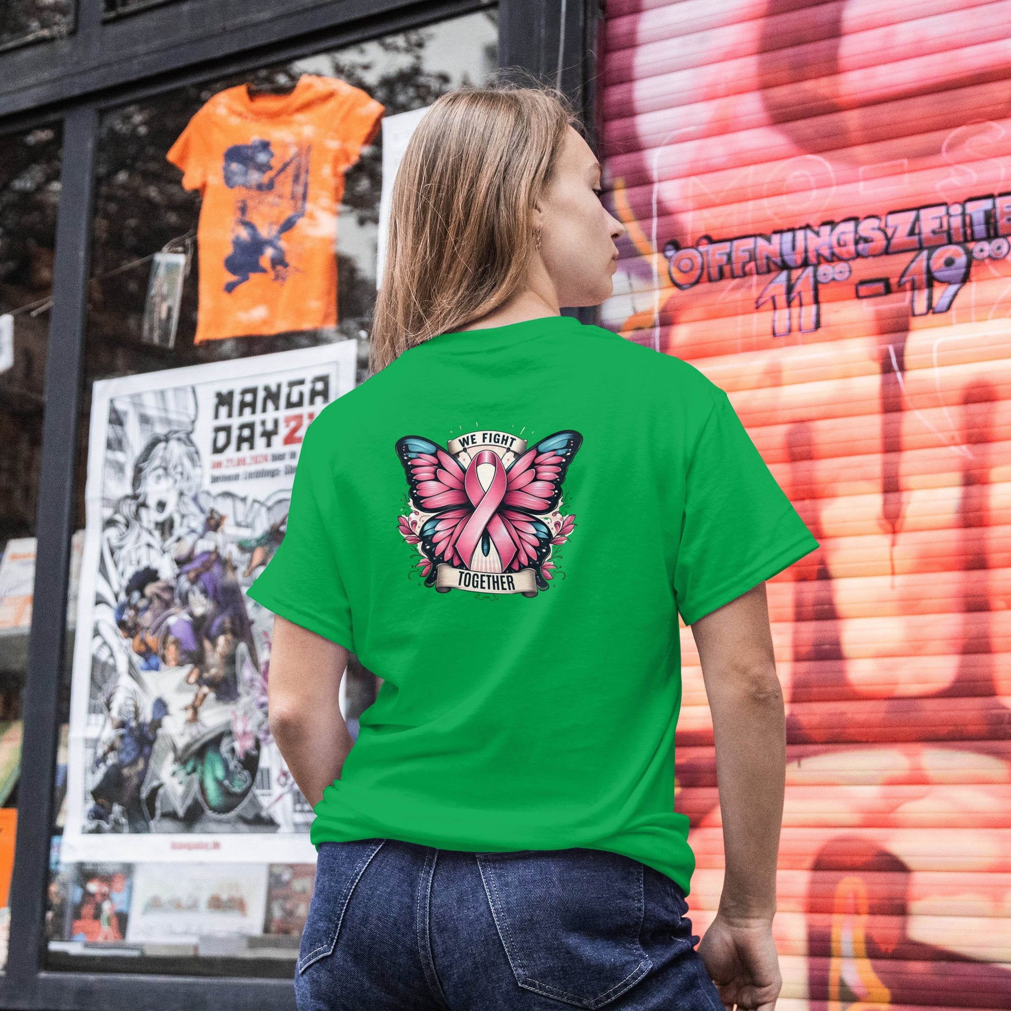 Person wearing a green t-shirt with a pink butterfly design and text, standing in front of a store window with posters and a colorful display.
