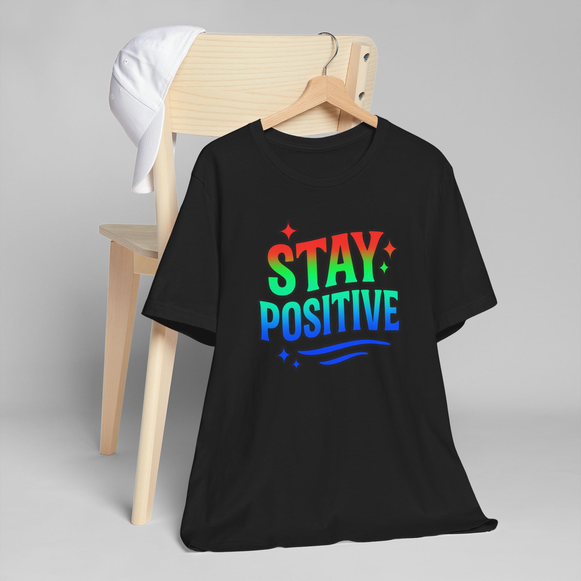 Black T-shirt with Stay Positive rainbow design on a wooden hanger, beside a white cap.