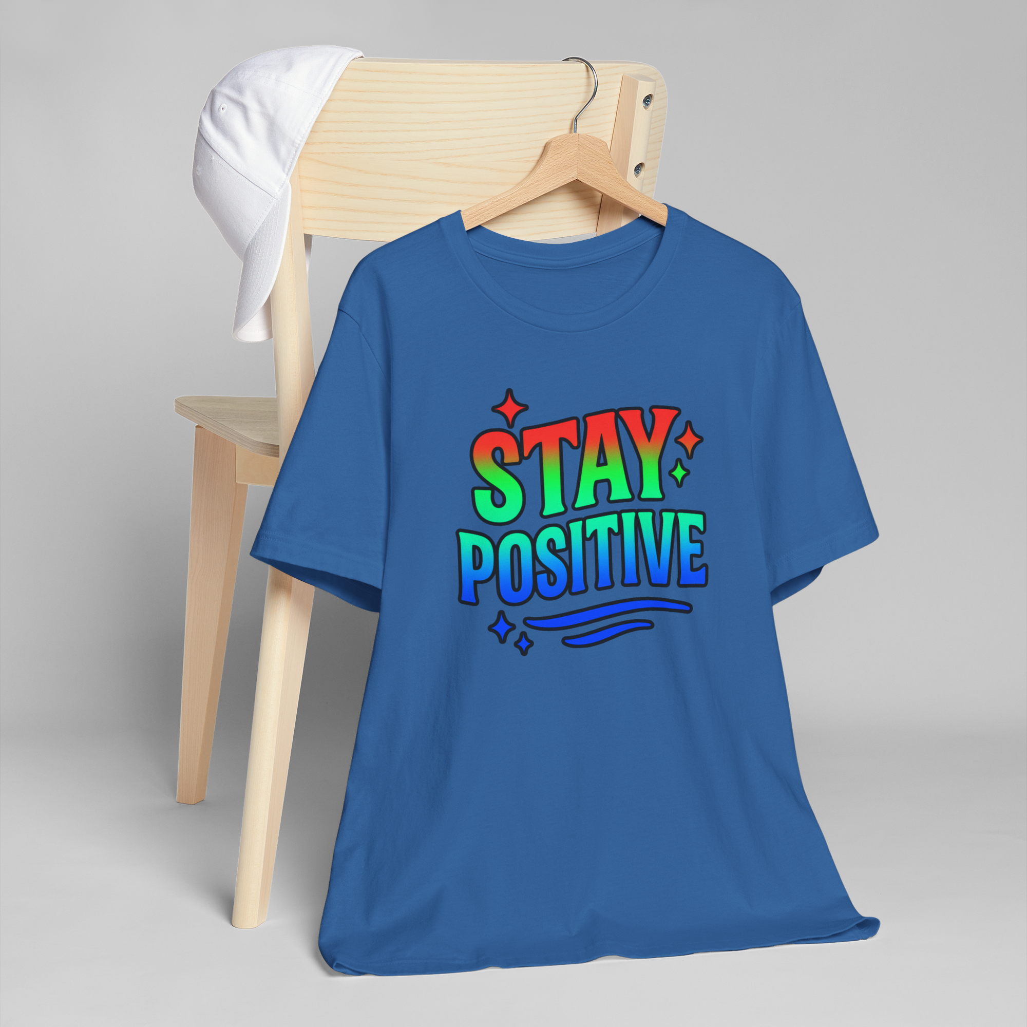 Blue Stay Positive T-shirt with rainbow gradient text, stars, and waves on a wooden hanger.