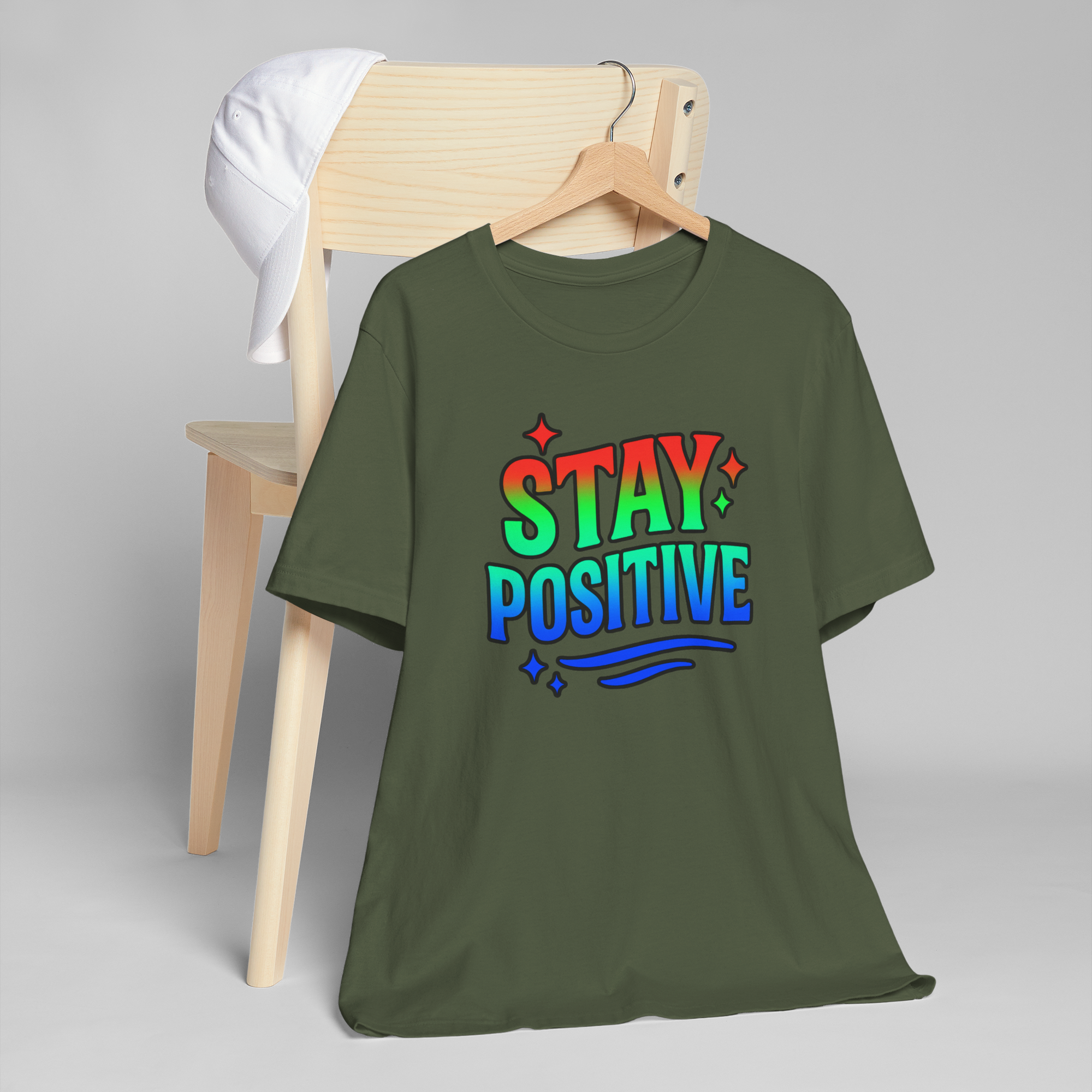 Olive green T-shirt featuring a rainbow Stay Positive graphic on a hanger, draped over a wooden chair with a white cap.
