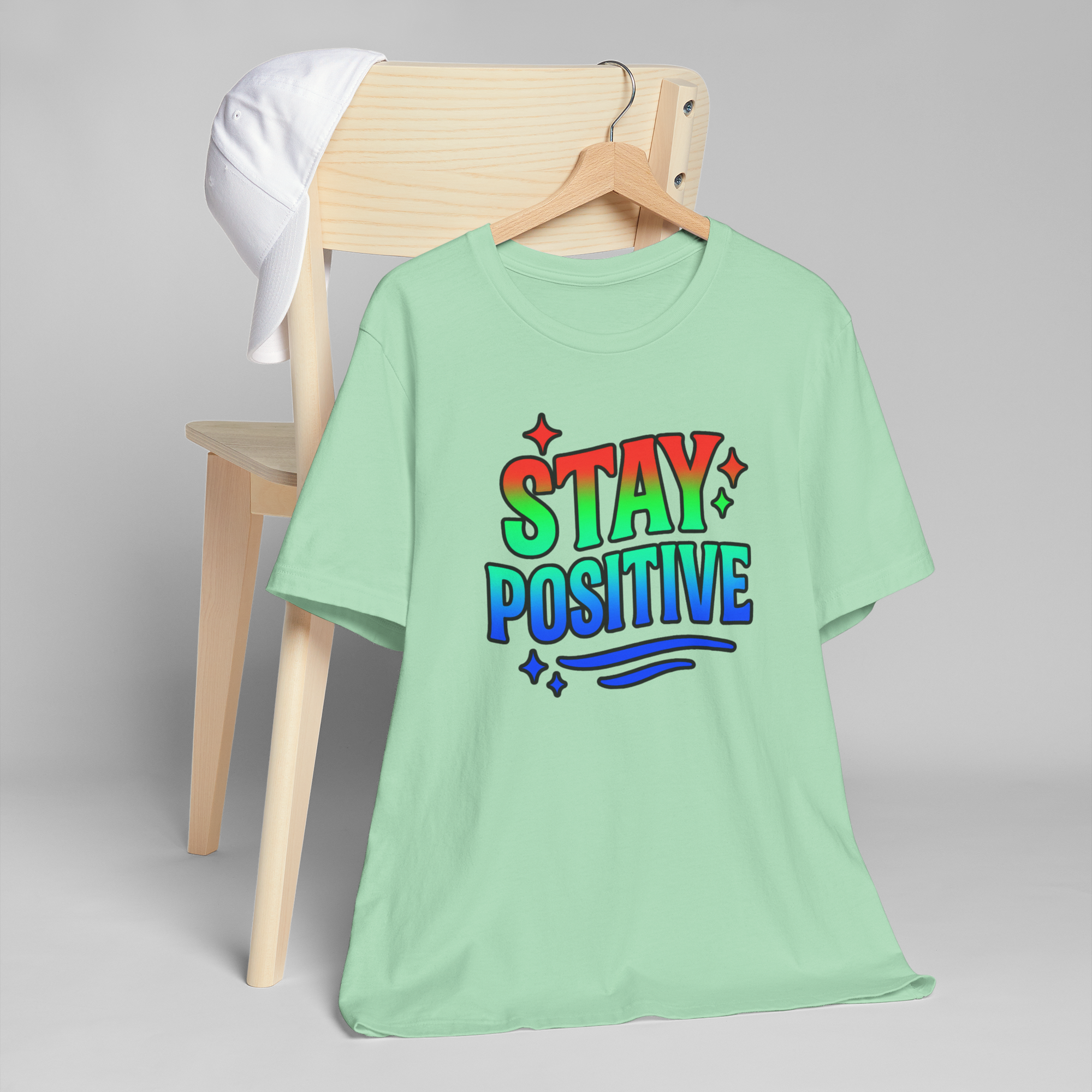 Mint green T-shirt on a wooden hanger with Stay Positive in rainbow gradient and stars; white cap on a chair.