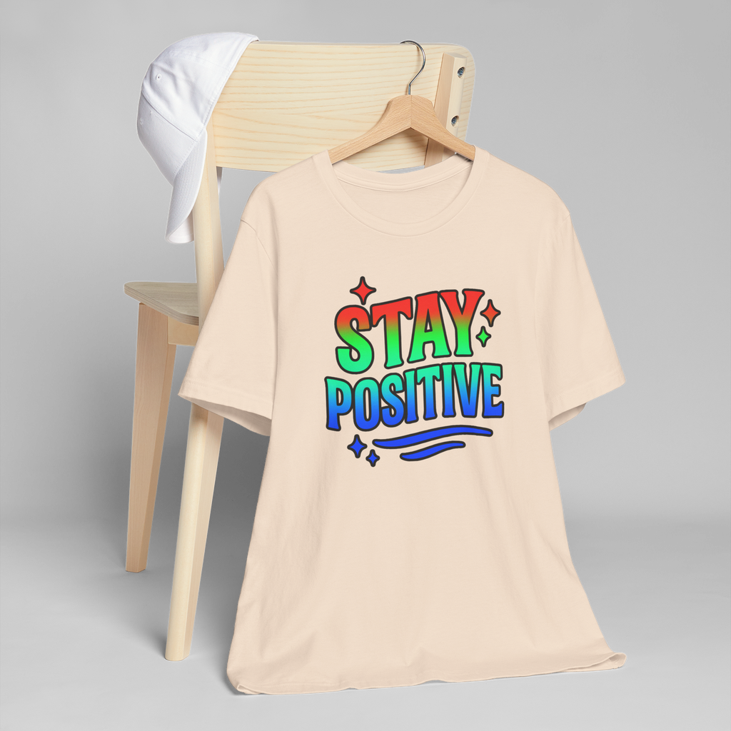 Stay Positive tee with rainbow gradient lettering and stars on a beige shirt hanging from a wooden chair and white cap.
