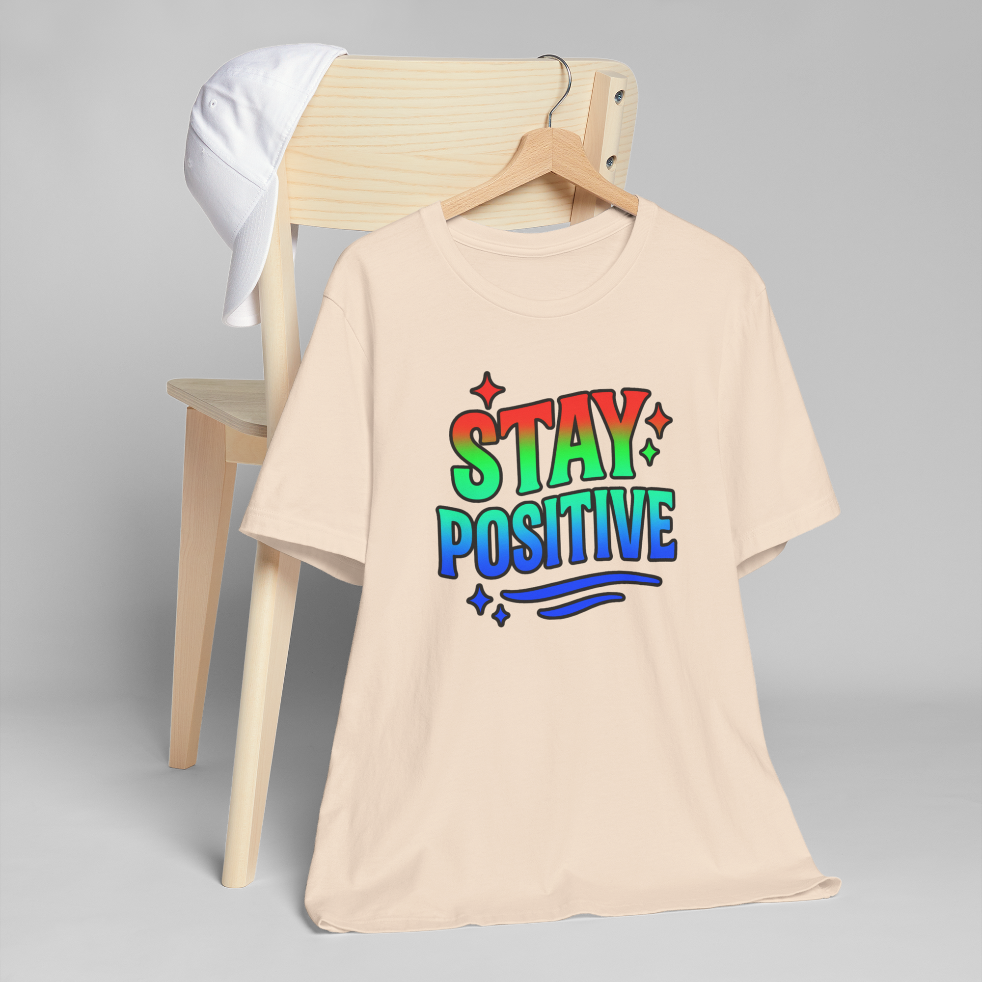 Stay Positive tee with rainbow gradient lettering and stars on a beige shirt hanging from a wooden chair and white cap.