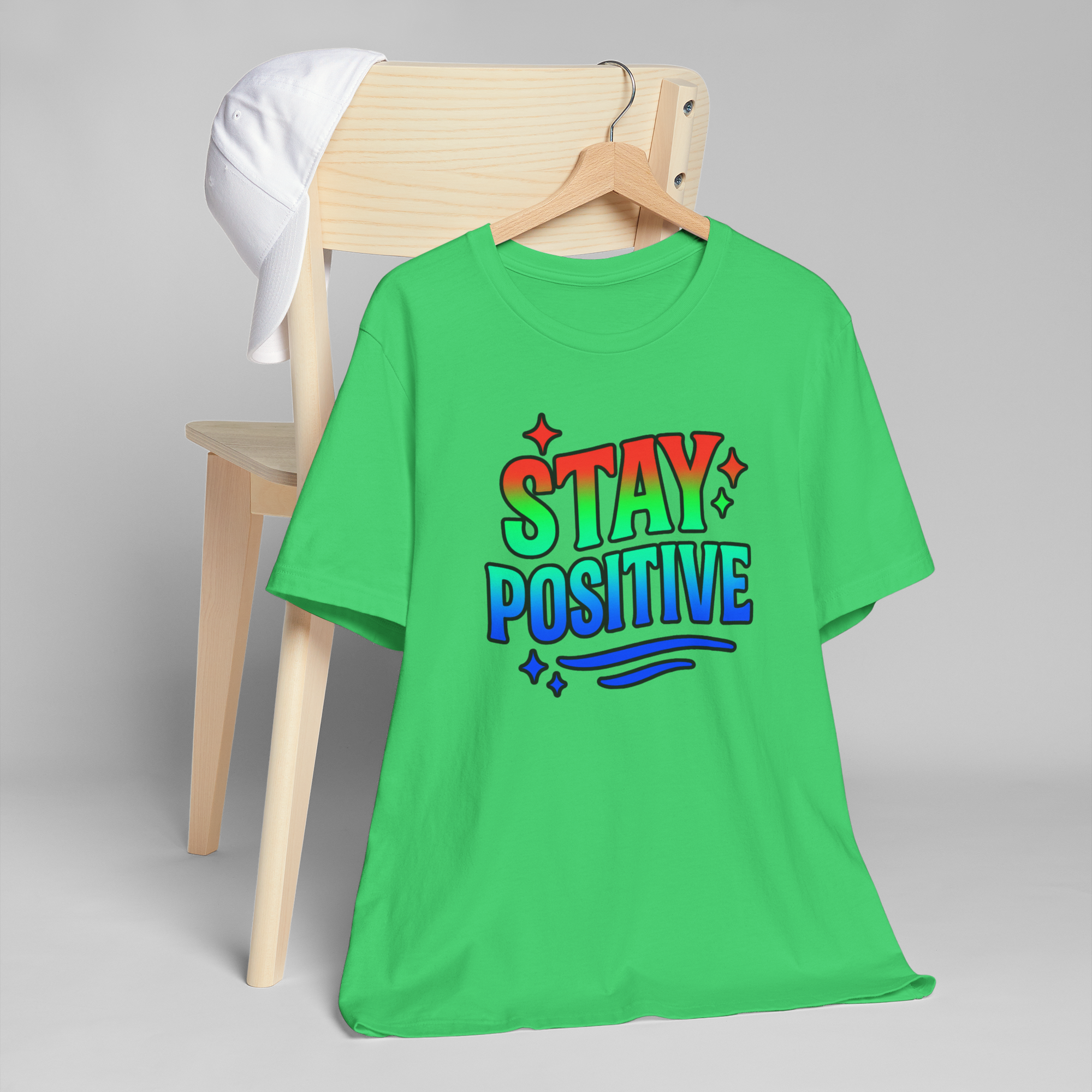 Green Stay Positive T-shirt with red-to-blue gradient text on hanger, white cap nearby.