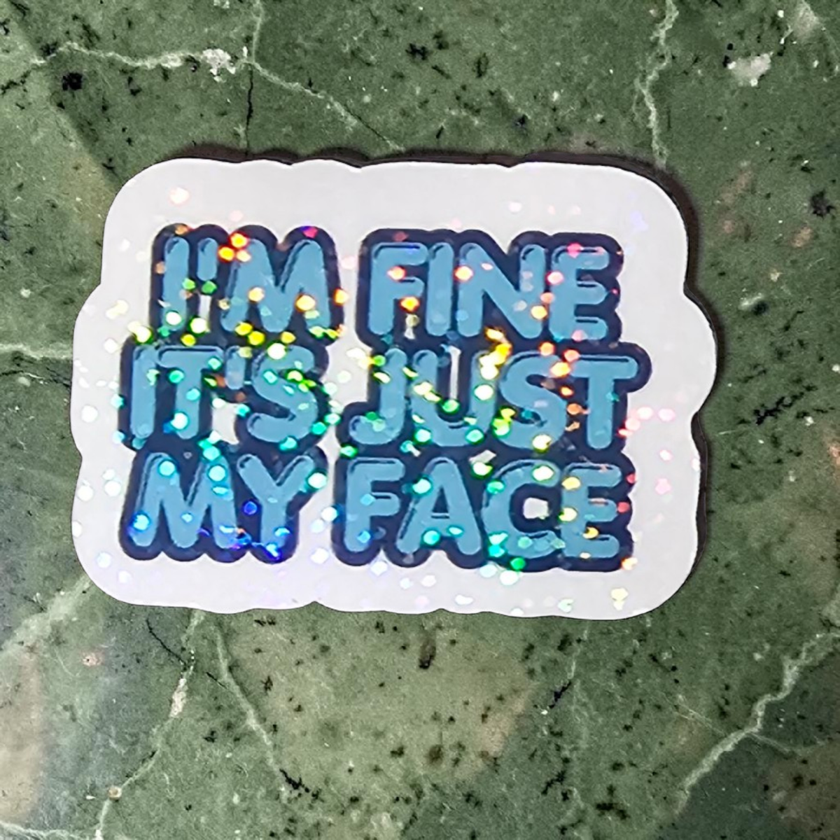Sticker with 'I'm fine it's just my face' text on a green surface