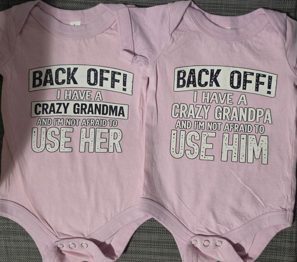 Two light pink baby bodysuits laid flat side by side on a dark textured surface. The left bodysuit reads 'BACK OFF! I HAVE A CRAZY GRANDMA AND I'M NOT AFRAID TO USE HER' and the right bodysuit reads 'BACK OFF! I HAVE A CRAZY GRANDPA AND I'M NOT AFRAID TO USE HIM'.