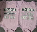 Two light pink baby bodysuits laid flat side by side on a dark textured surface. The left bodysuit reads 'BACK OFF! I HAVE A CRAZY GRANDMA AND I'M NOT AFRAID TO USE HER' and the right bodysuit reads 'BACK OFF! I HAVE A CRAZY GRANDPA AND I'M NOT AFRAID TO USE HIM'.