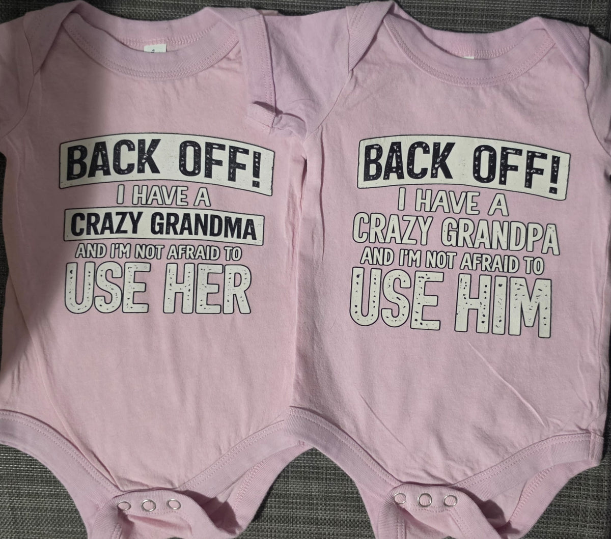 Two light pink baby bodysuits laid flat side by side on a dark textured surface. The left bodysuit reads 'BACK OFF! I HAVE A CRAZY GRANDMA AND I'M NOT AFRAID TO USE HER' and the right bodysuit reads 'BACK OFF! I HAVE A CRAZY GRANDPA AND I'M NOT AFRAID TO USE HIM'.