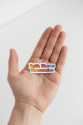Hand holding a colorful 'Faith Moves Mountains' sticker on a white background