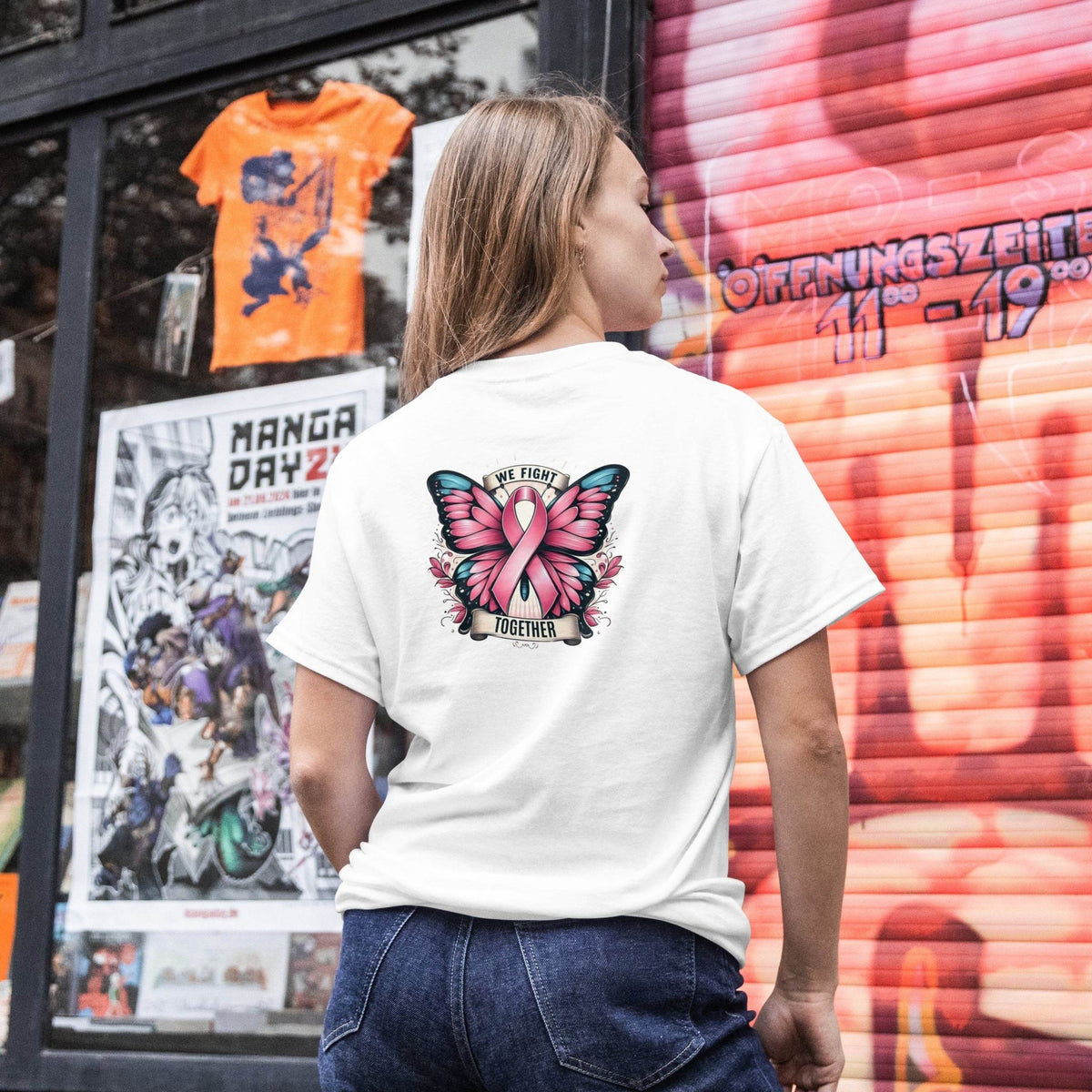 Person wearing a white t-shirt with a butterfly design and pink ribbon, standing in front of a colorful store window.