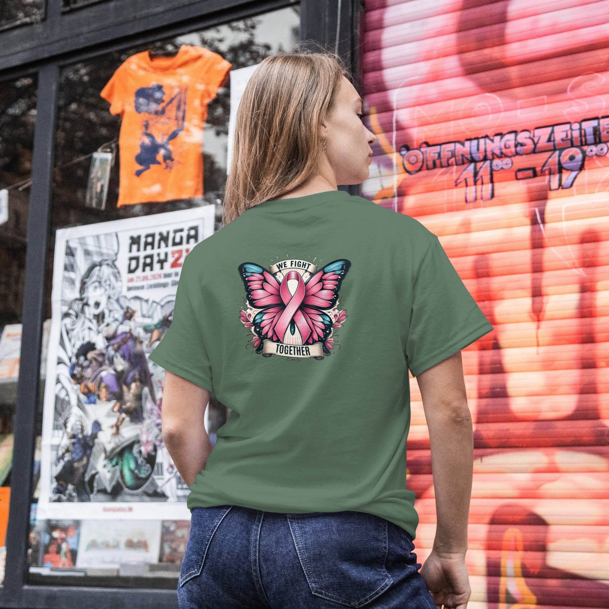 Person wearing a green t-shirt with a pink butterfly design and text, standing in front of a store window with posters and a colorful wall.