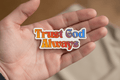 Hand holding a colorful sticker with 'Trust God Always' text against a blurred background
